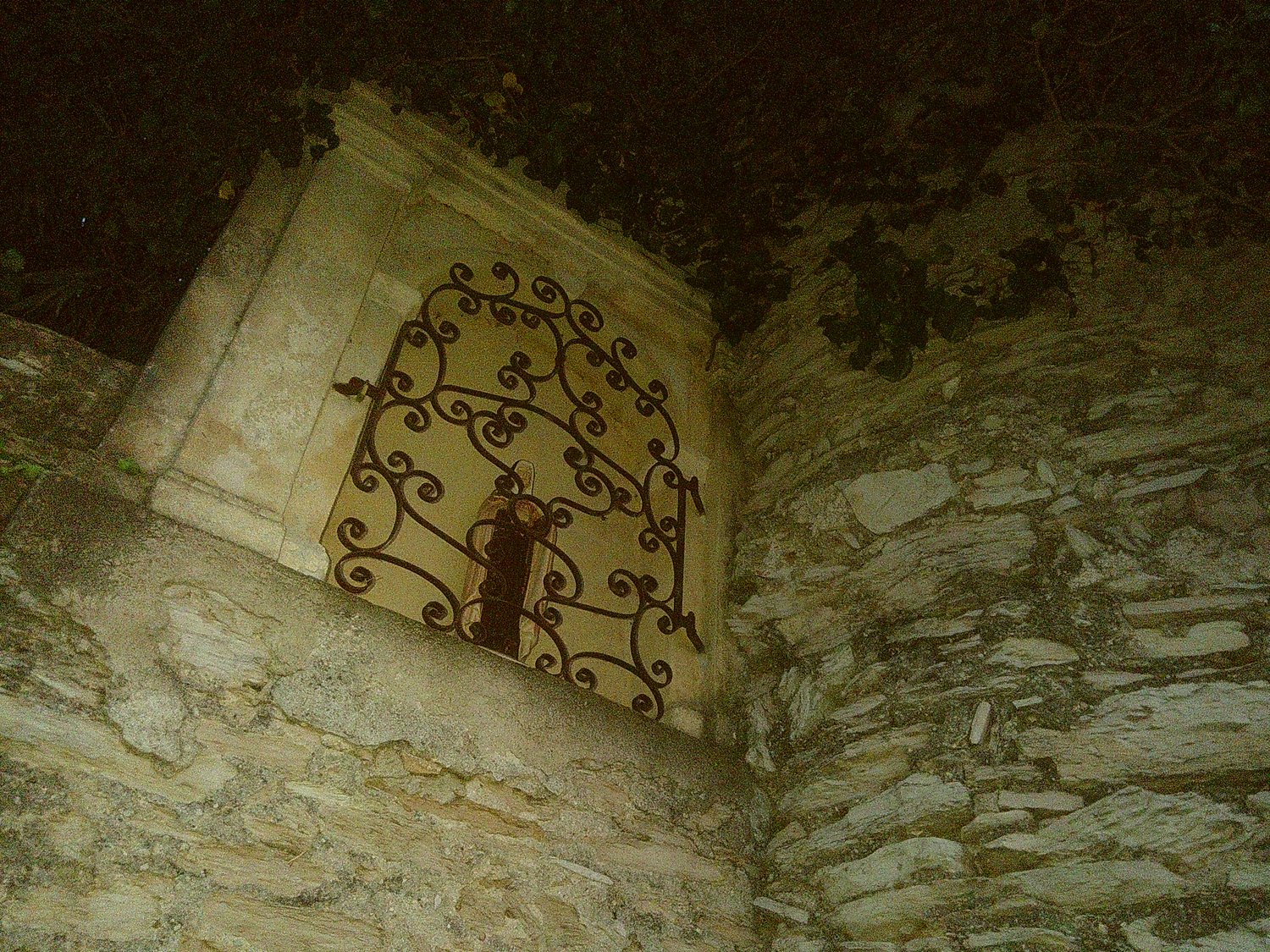 A corner in an old stone wall. A small chapel with an icon statue behind a metal grid door.